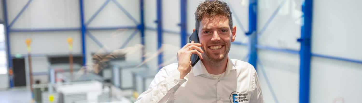 Een man in een wit shirt staat binnen, glimlachend en pratend op een telefoon. Hij staat voor een groot raam met uitzicht op industriële apparatuur en machines in een goed verlichte faciliteit.