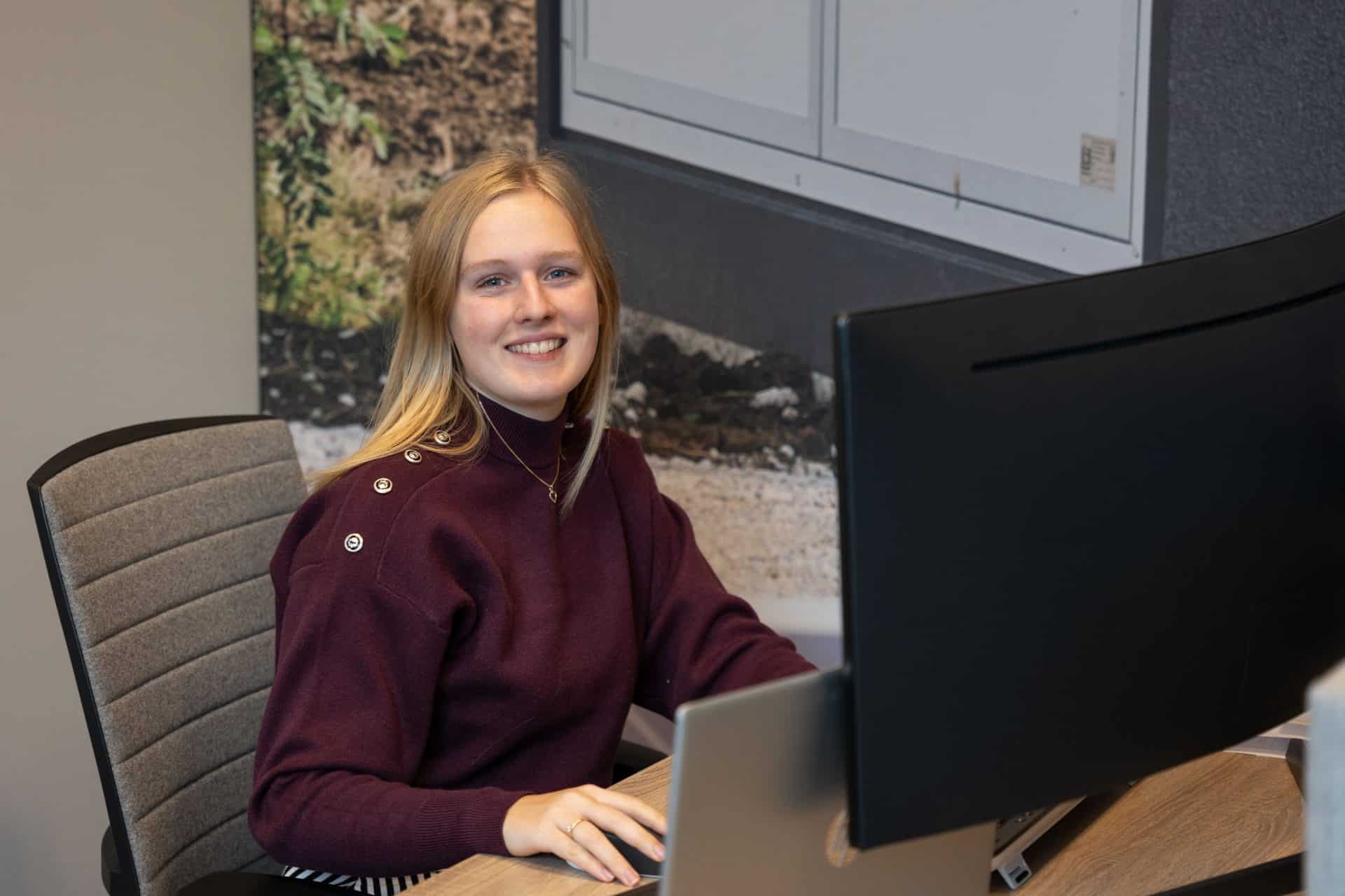 Een vrouw met lang blond haar, gekleed in een kastanjebruine trui, zit aan een bureau en werkt op een computer. Ze lacht naar de camera.