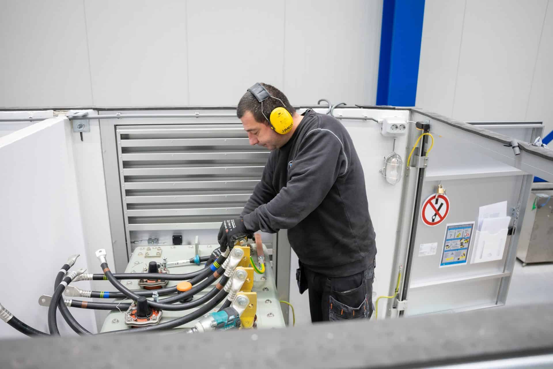 Een man met gele gehoorbescherming en handschoenen werkt in een fabriek aan industriële machines met verschillende slangen en onderdelen.