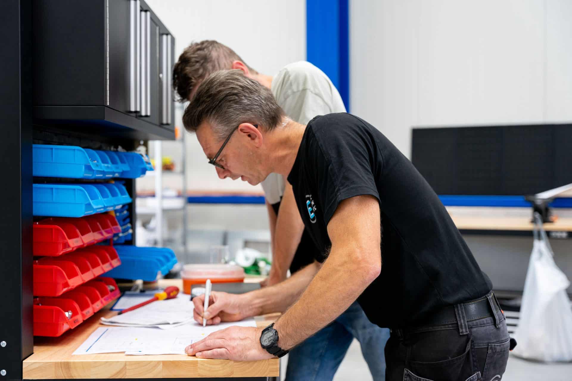 Twee mannen werken samen aan een werkstation, de een schrijft op papier terwijl de ander observeert. Het werkstation heeft georganiseerde opbergbakken met verschillende items.