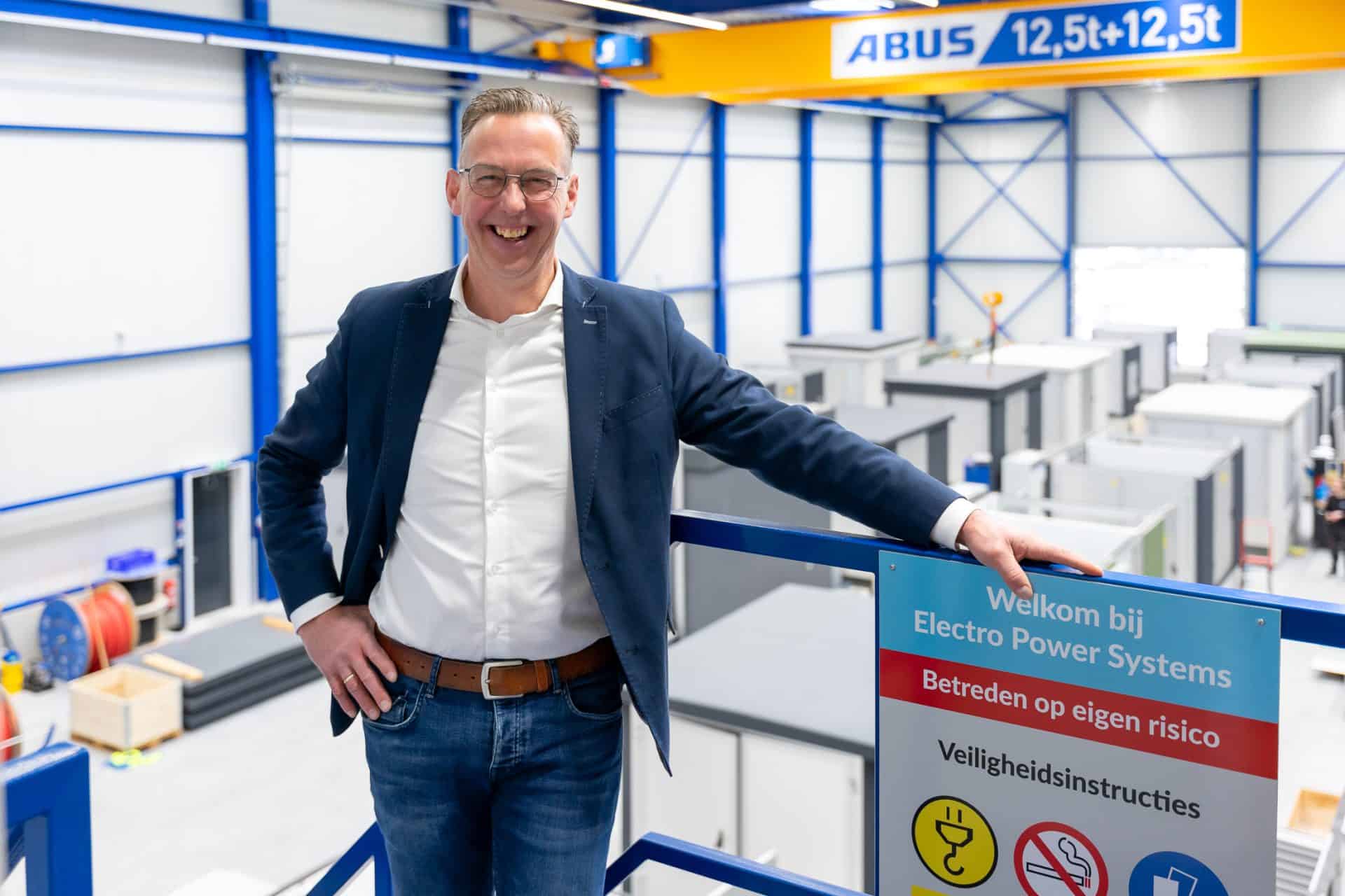 Een man in een blauw jasje en jeans staat lachend naast een bord met de tekst 'Welkom bij Electro Power Systems'. Op de achtergrond is een industriële omgeving met apparatuur en machines te zien.