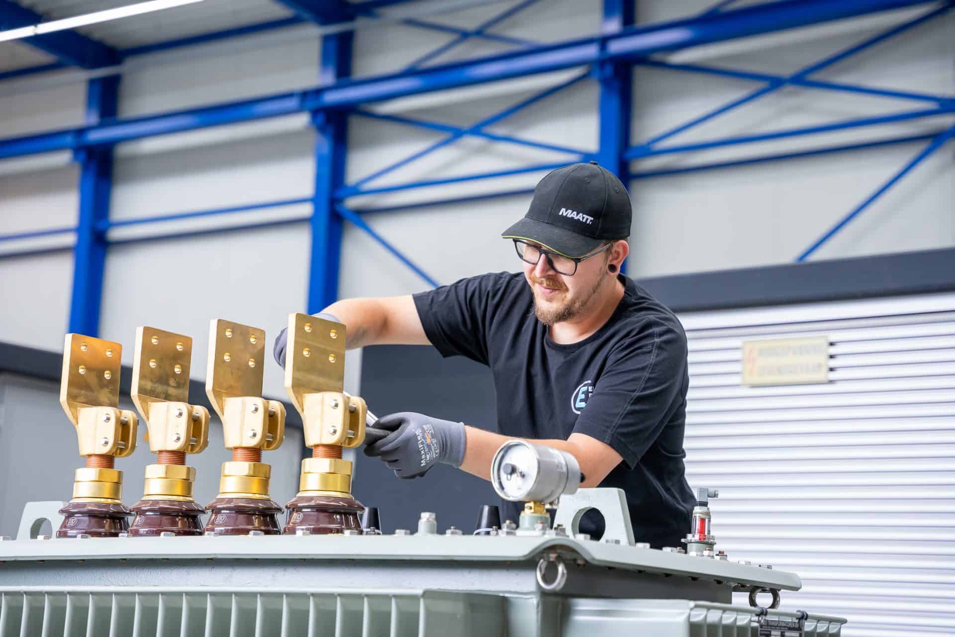 Een persoon met een zwarte muts en handschoenen werkt in een fabriek aan een industrieel apparaat met vier koperkleurige componenten.