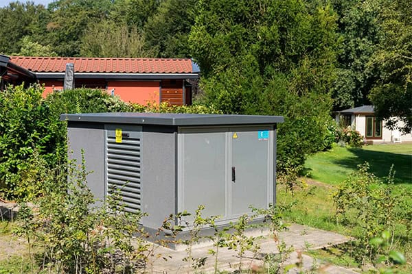 Een kleine, grijze utility box van Electro Power Systems staat buiten op een betonnen plaat in een tuin, omringd door struiken en bomen. Op de achtergrond zorgen rode en witte gebouwen voor een levendig contrast.