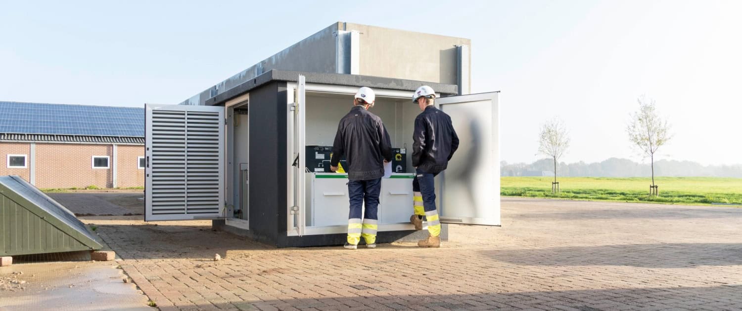 Twee middenspanningsmonteurs in beschermende kleding inspecteren apparatuur in een grote elektrische buitenkast op een verhard terrein bij een grasveld.