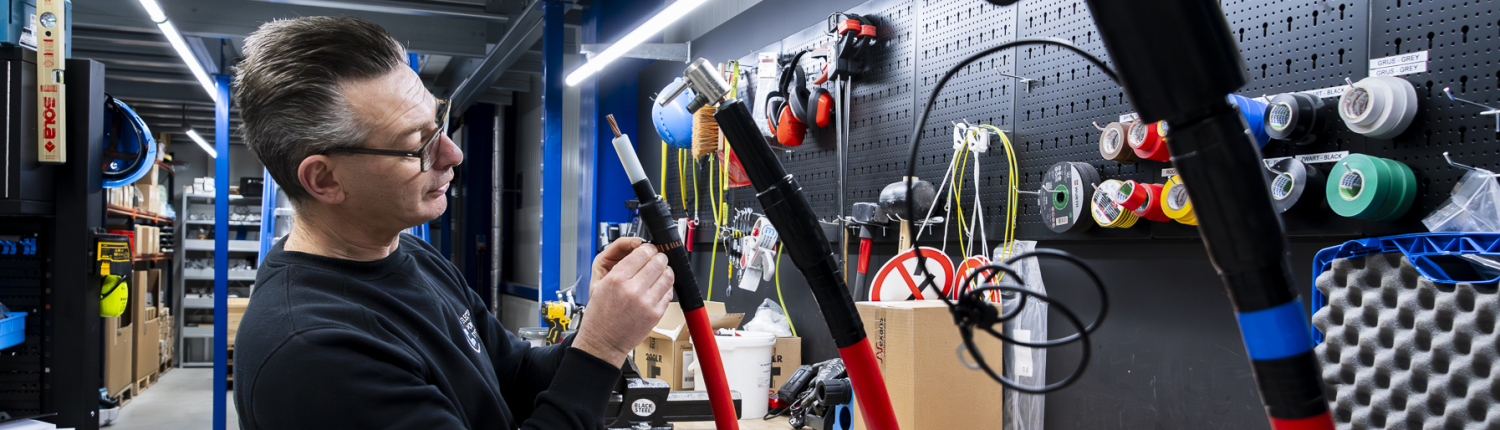 Een middenspanningsmonteur in een werkplaats inspecteert apparatuur met rode buizen, omringd door diverse gereedschappen en een overzichtelijk geperforeerd prikbord.