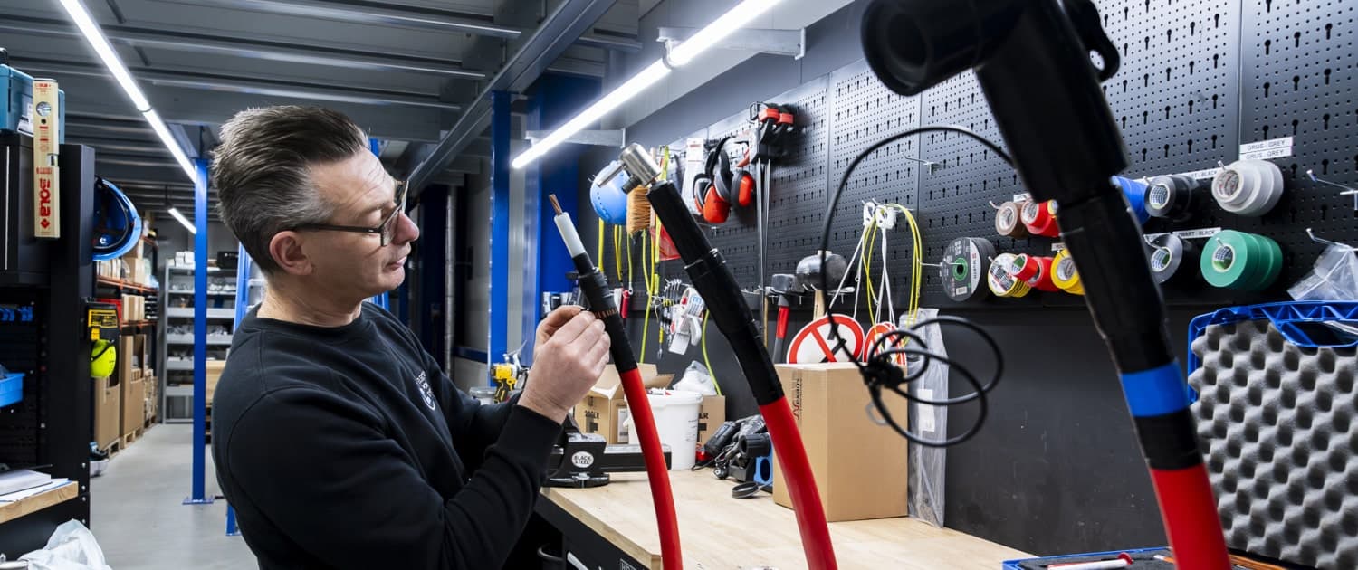 Een middenspanningsmonteur in een werkplaats inspecteert apparatuur met rode buizen, omringd door diverse gereedschappen en een overzichtelijk geperforeerd prikbord.