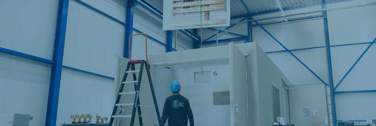 In een grote industriële faciliteit staat een arbeider met een blauwe helm naast een container, met een ladder en elektrische apparatuur in de buurt. Deze momentopname van Nieuwsbericht oktober 2024 benadrukt de ingewikkelde dans van industrie en veiligheid.